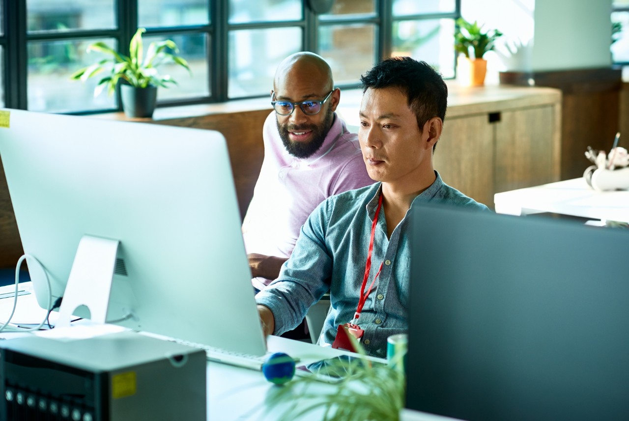 Mid adult Chinese man sitting at desk using computer, IT supporter assisting office worker, software developer looking at screen and concentrating