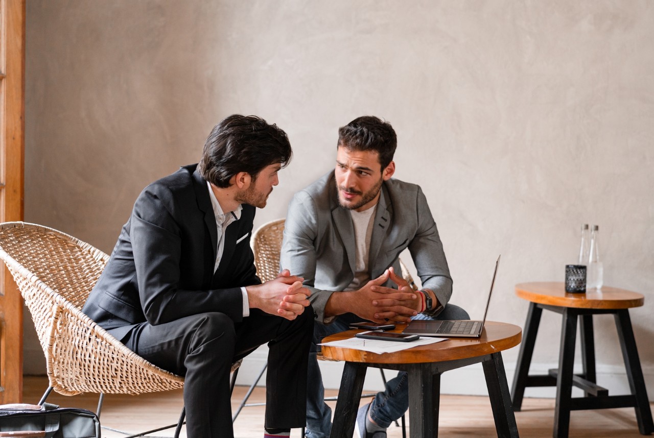 Two coworkers are having a meeting in an open office environment. They are discussing around their laptop and some documents.